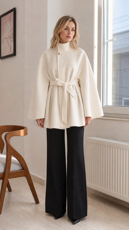 Front view of woman wearing cream high-neck belted cape coat with black trousers