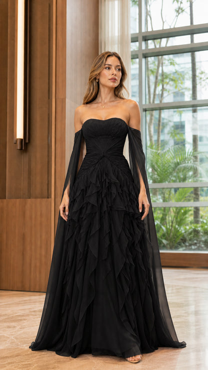 Model wearing black strapless tiered ruffle evening gown with draped cape sleeves