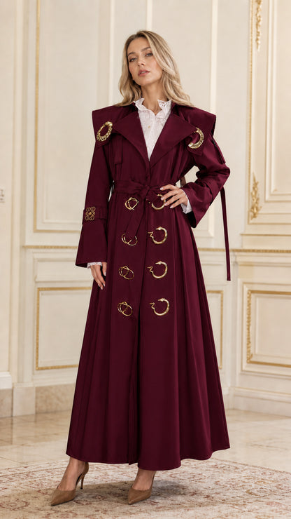 Front view of woman wearing burgundy maxi belted trench coat with gold accents